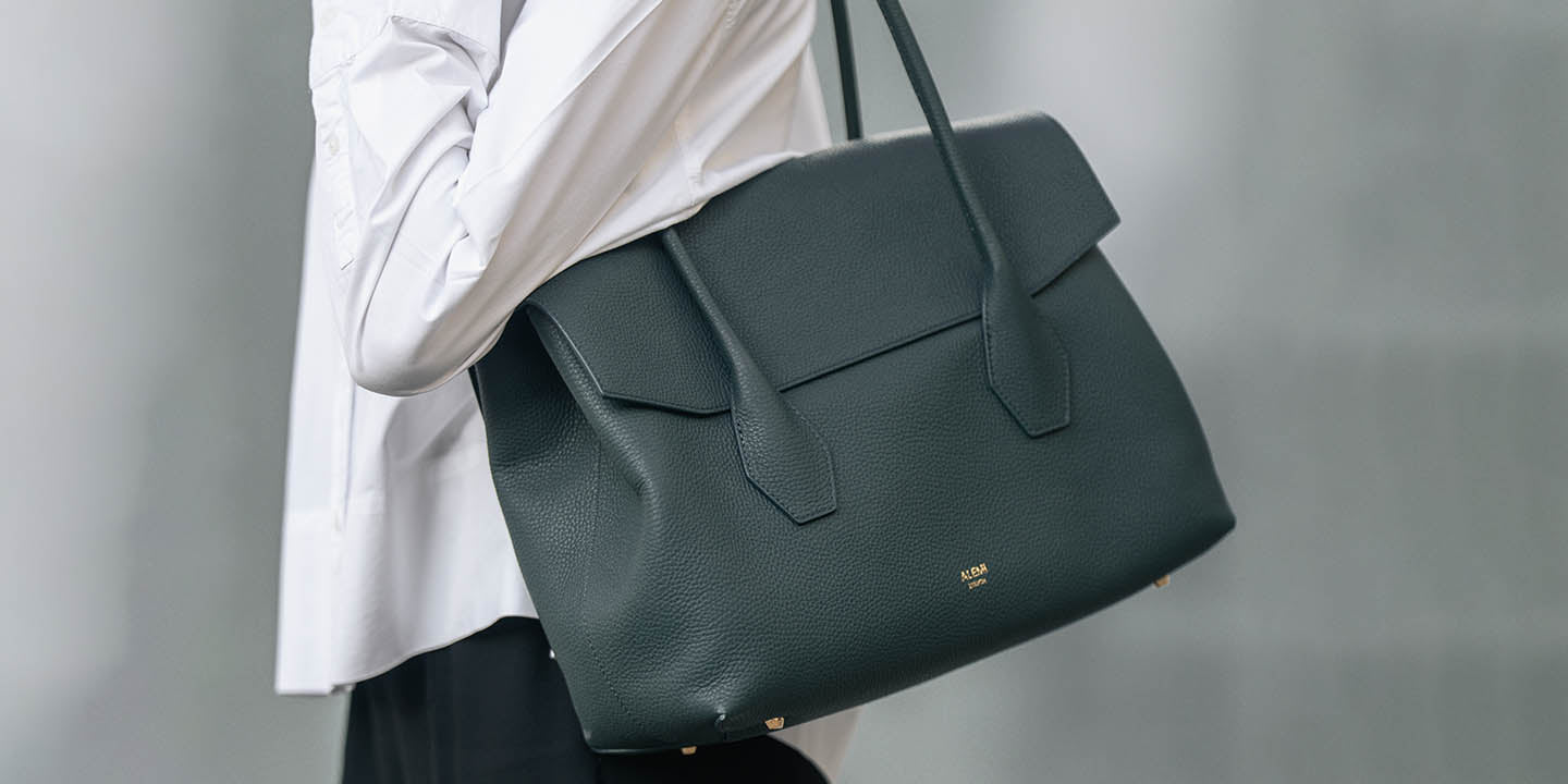 A woman in a white shirt and black pants carries a large deep teal pebbled leather IDA Bag from ALEMI with gold-tone hardware against a soft gray background.