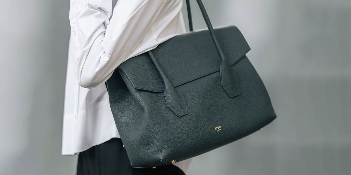 A woman in a white shirt and black pants carries a large deep teal pebbled leather IDA Bag from ALEMI with gold-tone hardware against a soft gray background.